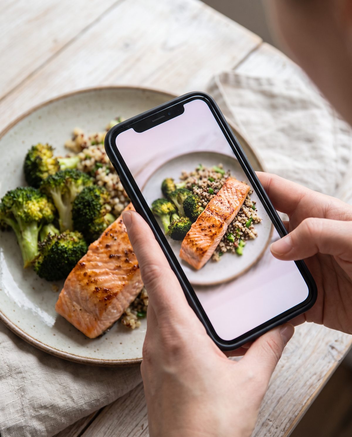Hands holding a phone over a plate of salmon, broccoli and quinoa